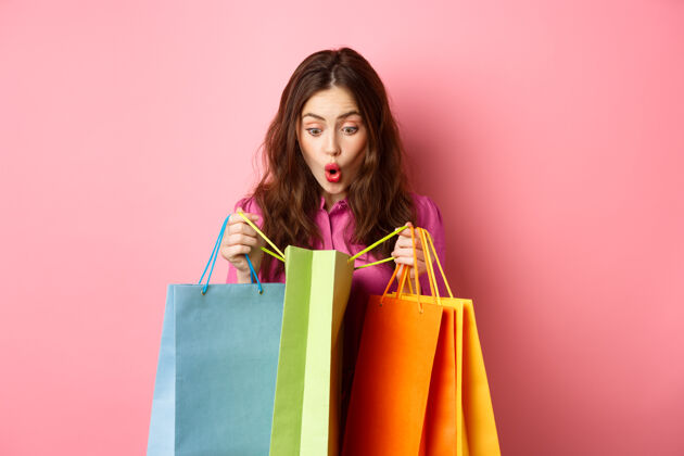请兴奋的女孩打开购物袋 大吃一惊 兴高采烈地为她挑选礼物 站在粉红色的墙上复制空间女性购物成人