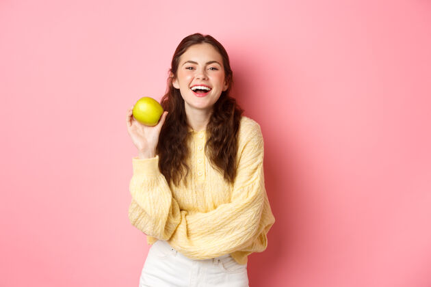 惊人健康的饮食 人和生活方式的概念苹果日远离医生 女孩拿着美味的水果 微笑着对着镜头 站在粉红色的墙上时尚魅力微笑