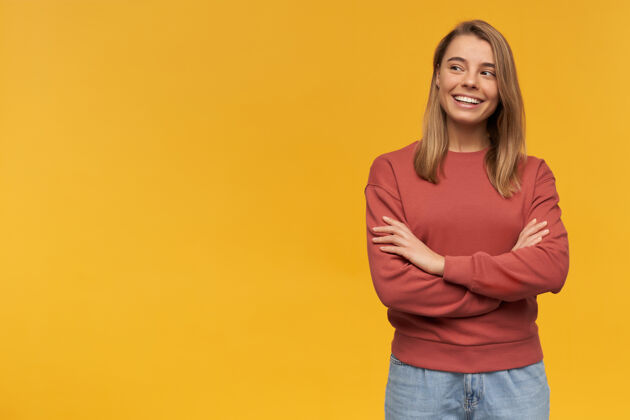 女人穿着陶土运动衫的快乐美丽的年轻女子站在一旁 双手交叉放在黄色的墙上高兴女性青少年