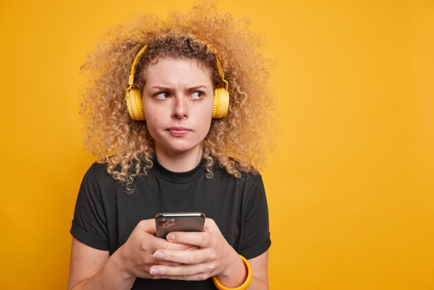思考嚴重不高興的卷發女子使用免費上網連接在線聽音樂戴著無線耳機戴在耳朵上穿著休閑的黑色t恤在室內擺出體貼的表情姿勢互聯網成人手持