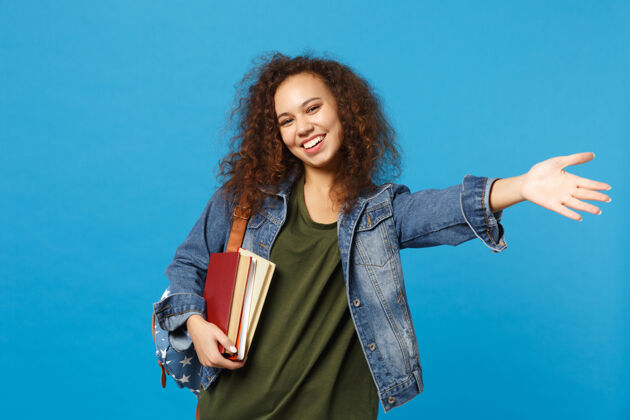 学习穿着牛仔服 背着书包的年轻女学生把书孤立地放在蓝色的墙上快乐学者背包