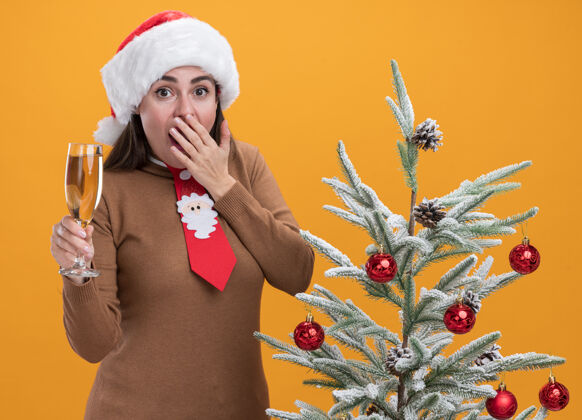 抱着被吓得戴着圣诞帽打着领带的年轻漂亮女孩站在圣诞树旁 手里拿着一杯香槟酒 嘴巴蒙着 手被隔离在橙色的墙上帽子掩护女孩
