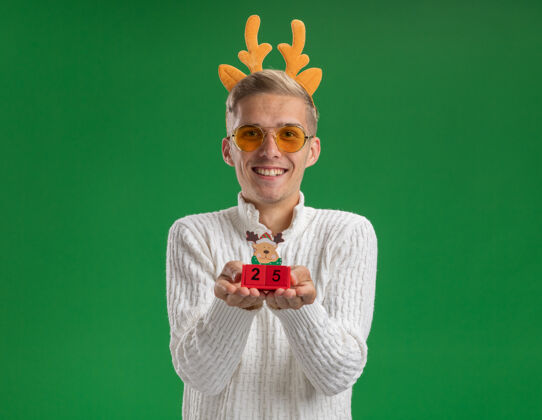 帅哥微笑的年轻帅哥戴着驯鹿鹿角头带戴着眼镜拿着圣诞树玩具和日期看起来孤立的绿色墙壁上的复制空间约会驯鹿年轻