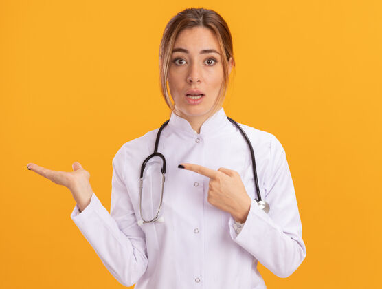 姿势困惑的年轻女医生穿着医用长袍 听诊器假装拿着 指着黄色墙上孤立的东西穿着人表情