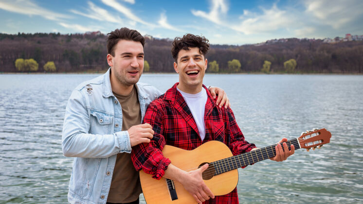 阳光两个年轻的朋友在公园的湖边唱歌和弹吉他朋友团体放松