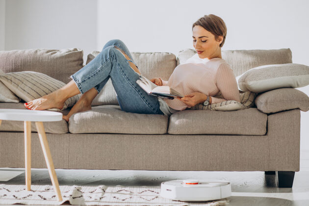 易拿年轻的女人躺在沙发上看书 而机器人吸尘器做家务机器家具沙发