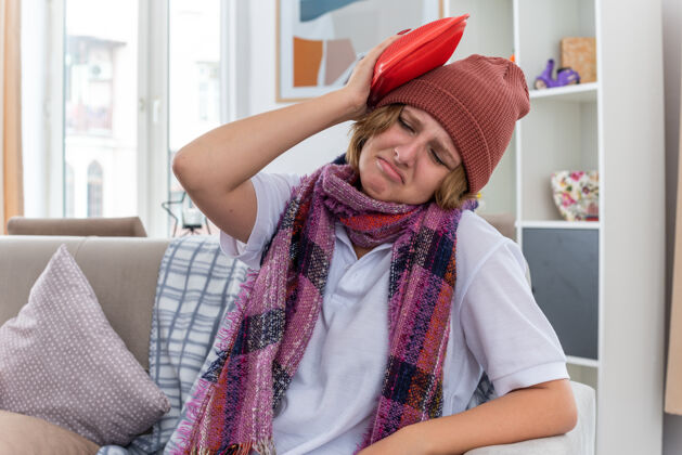 生病不健康的年轻女子戴着帽子 脖子上围着暖和的围巾 感觉不舒服 生病 患感冒和流感 头上戴着热水瓶 坐在客厅里的沙发上 看起来很担心沙发头瓶子