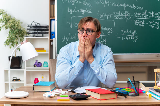 年轻年轻的男老师戴着眼镜看着一边紧张地捂着嘴坐在课桌旁 手里拿着书和笔记放在教室的黑板前压力穿放一边