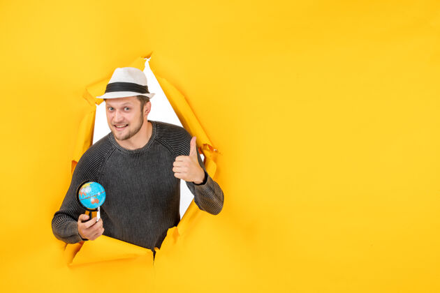 帽子戴着帽子的年轻人拿着一个小地球仪 在撕破的黄色墙壁上做着一个表情愉快的ok手势摩托板撕裂面部