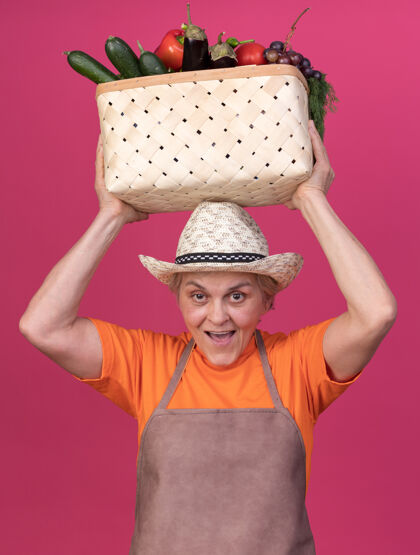 穿快乐的上了年纪的女园丁戴着园艺帽 头顶着菜篮子粉红色蔬菜举行