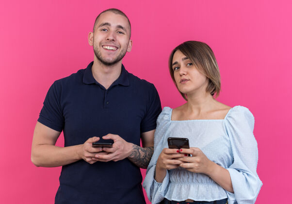 男人一对年轻漂亮的夫妇 带着智能手机 站在粉色的墙上开心地笑着站立年轻人欢呼