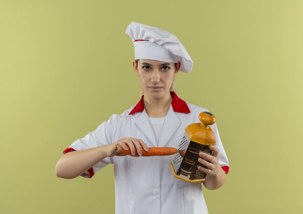 烹饪自信的年轻漂亮厨师身着厨师制服 用隔离在绿墙上的烤炉烤胡萝卜自信感激制服