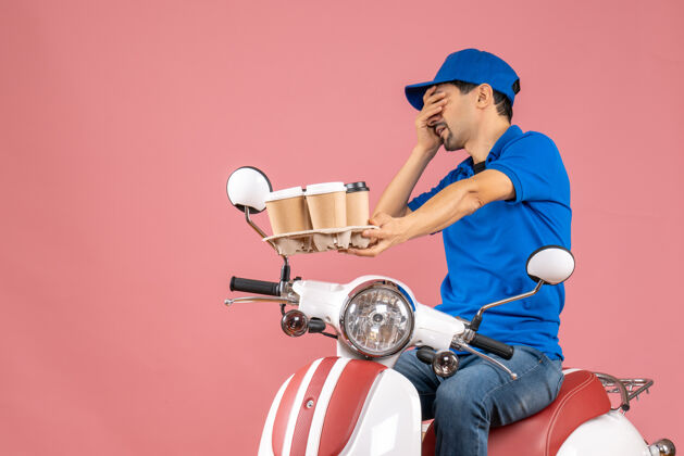 男疲惫的快递员戴着帽子坐在粉彩桃色背景上的滑板车上快递员坐桃