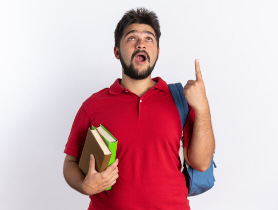 胡须年轻的留着胡子的学生 穿着红色马球衫 背着背包 拿着笔记本 高兴地抬头 兴奋地用食指指着白色的墙壁人背包手指