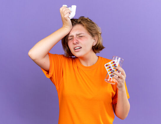 感觉不健康的年轻女子穿着橙色t恤 站在紫色的墙上 手里拿着一杯水和药片 摸着她的额头 因为生病 发烧 流感和感冒病毒触摸发烧药丸