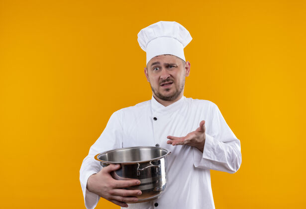 手困惑的年轻帅哥厨师穿着厨师制服拿着锅炉 在孤零零的橘色墙上空手展示锅炉烹饪困惑