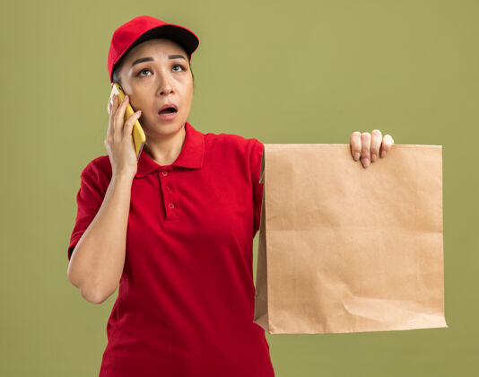 惊喜年轻的送货员身穿红色制服 戴着帽子 手里拿着一个纸包裹 站在绿色的墙上 一边用手机交谈 一边显得困惑和惊讶拿着帽子年轻