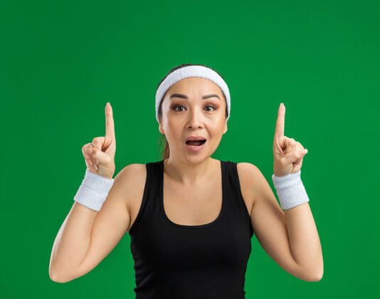 女人戴着头带和臂章的年轻健身女士惊讶地伸出食指站在绿色的墙上健身头带年轻