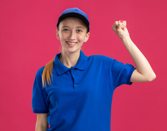 帽子身着蓝色制服 头戴鸭舌帽的快乐少女站在粉红色的墙上自信地举起拳头提高信心微笑