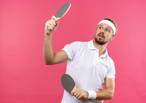 印象印象深刻的年轻英俊的运动男子戴着头带和腕带举行乒乓球拍 看着他们在粉红色的墙壁与复制空间孤立帅气头带年轻