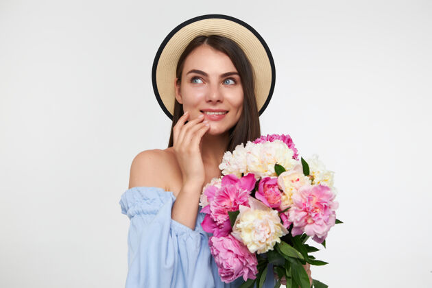 室内一个快乐的女人 深色的长发 戴着帽子 穿着蓝色的裙子 手里拿着一束花 抚摸着下巴头发梦女士
