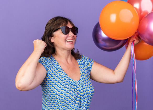 立场快乐和兴奋的中年妇女手持一束五颜六色的气球握紧拳头庆祝生日聚会站在紫色的墙上握紧庆祝生日
