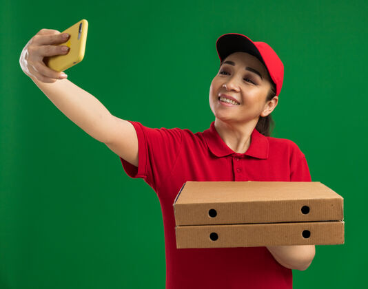 站着身穿红色制服 头戴鸭舌帽 手持披萨盒 用智能手机自拍的年轻送货员站在绿色的墙壁上开心地微笑着披萨女人帽子