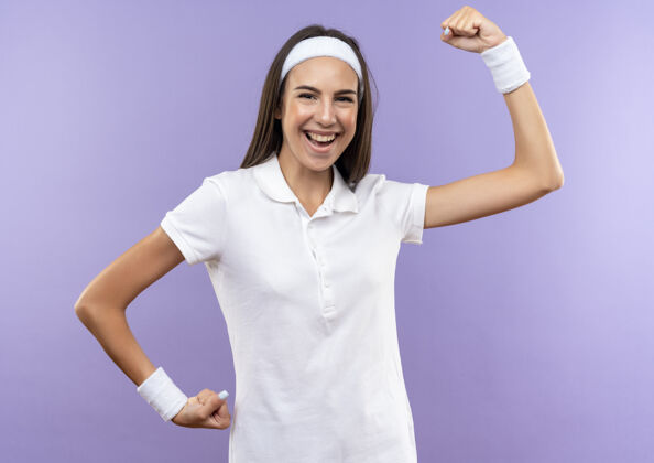 强壮快乐漂亮的运动女孩戴着头带和腕带在紫色的墙上做着强烈的孤立的手势穿着头带快乐