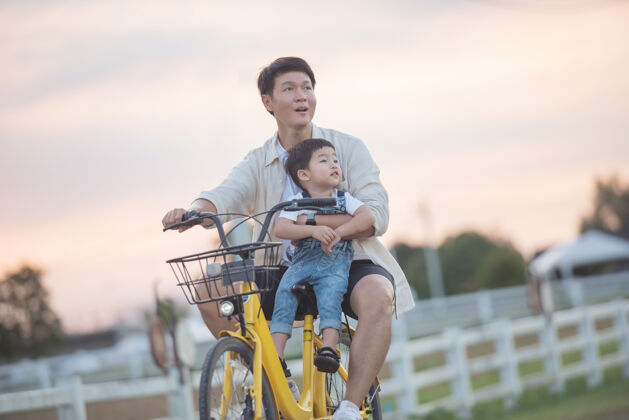 家庭一对骑着自行车的快乐的年轻父亲和儿子的画像日落时分 父亲和儿子在公园里玩耍人们在球场上玩耍友好家庭和暑假的概念儿子自行车男性