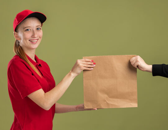 微笑年轻的送货员穿着红色制服 戴着帽子 手里拿着一个纸包 在绿色的墙壁上自信地微笑着送给顾客年轻包装女孩