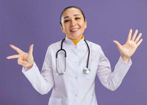 女人年轻的女医生 身穿白色药衣 脖子上戴着听诊器 脸上带着微笑 八号手指站在紫色的墙上年轻人外套听诊器