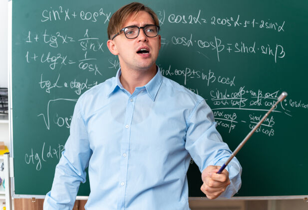 年轻愤怒的年轻男老师戴着眼镜 拿着指针 站在教室里数学公式的黑板旁讲解功课黑板公式靠近