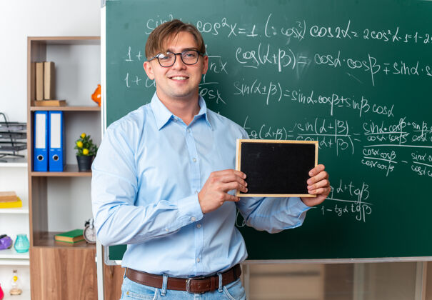 数学年轻的男老师戴着眼镜 拿着小黑板 面带微笑 自信地站在教室里的数学公式黑板旁数学老师男