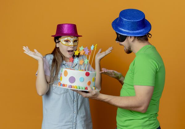面具快乐的帅哥戴着蓝色帽子戴着化装眼罩拿着生日蛋糕看着惊讶的年轻女孩戴着粉色帽子和化装眼罩举手看着蛋糕蓝色橙色蛋糕