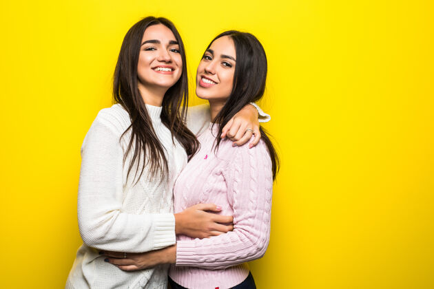 朋友两个穿着毛衣的快乐女孩在黄色墙壁上拥抱的肖像手指显示肖像