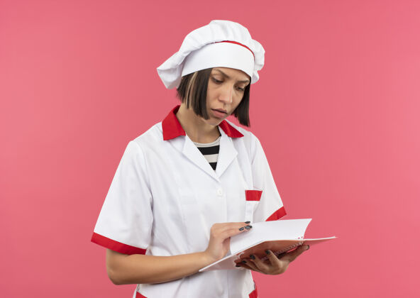 烹饪体贴的年轻女厨师 穿着厨师制服 拿着笔记本 看着隔离在粉红色墙上的笔记本人衣服制服