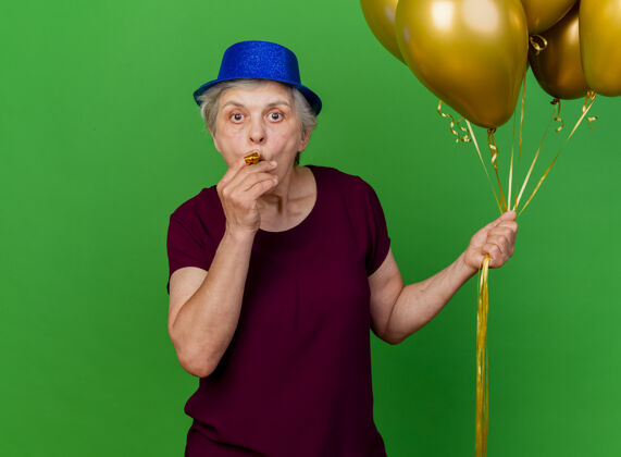 帽子惊讶的老妇人戴着聚会帽手持氦气球吹着绿色的口哨派对女人氦