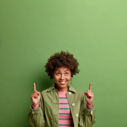 请开朗的女人指着上面的两个手指展示你的广告复制空间微笑着穿着时髦的衣服隔离在绿色的墙上年轻夹克学生