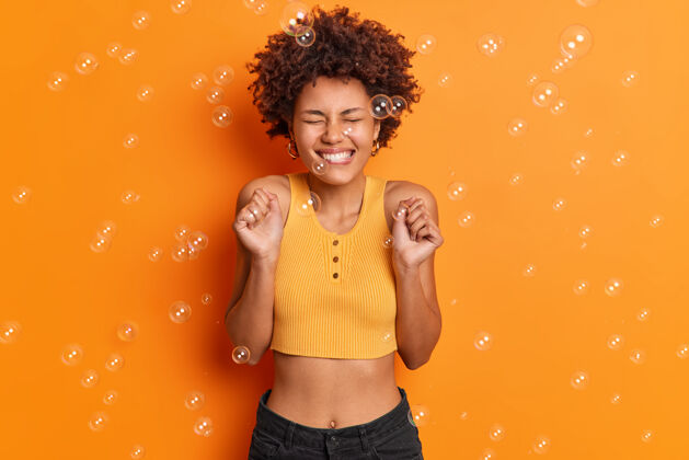 是快乐的胜利女人的水平镜头卷曲的非洲头发咬嘴唇高兴地得到批准或听到极好的消息穿着不规则的上衣和牛仔裤摆出反对橙色墙壁肥皂泡热情身体积极