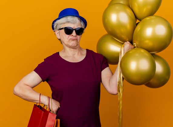 包戴着派对帽 戴着太阳眼镜 拿着氦气球和纸购物袋的老妇人气球氦聚会