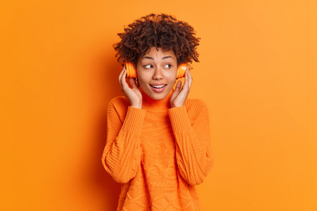 非洲心满意足的卷发女人通过耳机收听音轨看着一旁微笑愉快地穿着休闲毛衣隔着橙色的墙人享受高兴