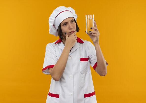 烹饪体贴的年轻女厨师 身着厨师制服 手放在橘黄色的墙上 举着一杯水 看着水衣服杯子年轻