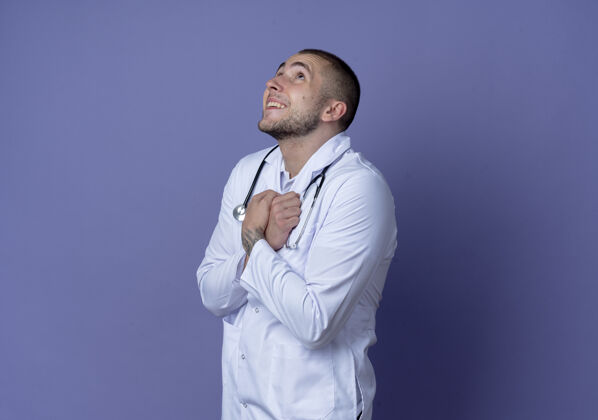 紫色面带微笑的年轻男医生穿着医用长袍 手持听诊器站在侧视图中 双手交叉放在胸前 孤立地看着紫色的墙上人穿着站立