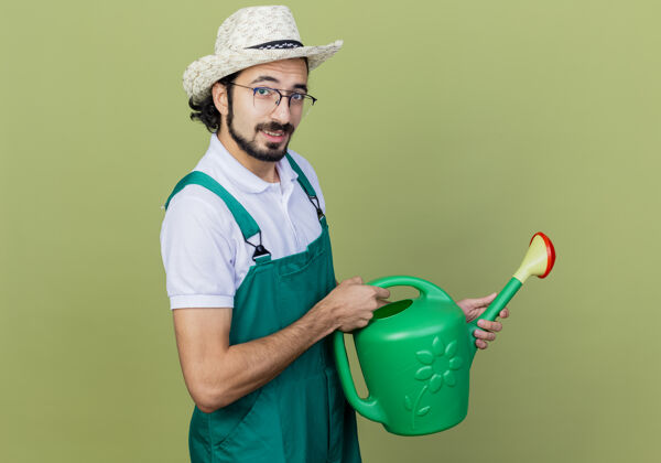感觉年轻的留着胡须的园丁 穿着连体衣 戴着帽子 拿着喷壶 站在浅绿的墙上微笑着看着前面人帽子抱着