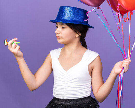 帽子戴着蓝色派对帽 手持氦气球 看着紫色墙壁上孤立的派对哨子 还有复制空间孤立气球氦