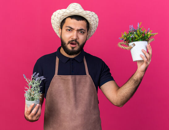 年轻未出租的年轻白人男性园丁戴着园艺帽拿着花盆隔离在粉红色的墙上复制空间持有男性不愉快