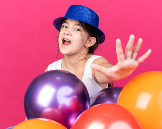 伸展焦急的年轻白人女孩戴着蓝色的聚会帽 站在氦气球旁 伸出一只孤立的手 站在粉色的墙上 留着复印空间氦帽子穿
