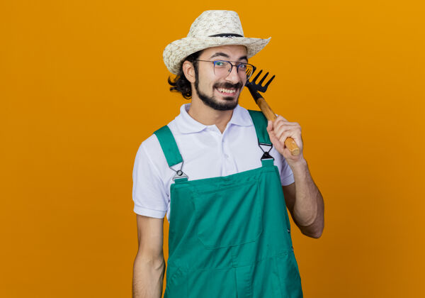 快乐年轻的留着胡须的园丁 穿着连体衣 戴着帽子 拿着小耙子 站在橘色的墙上 面带微笑地看着前面姿势帽子脸