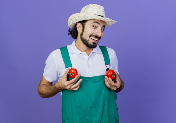 站着有趣的年轻留着胡子的园丁 穿着连体衣 戴着帽子 手里拿着新鲜的西红柿 站在蓝色的墙上 兴高采烈地微笑着看着前面人花园感觉