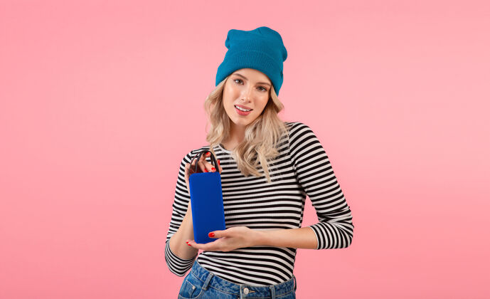 配件年轻女子手持无线扬声器听音乐穿着条纹衬衫和蓝色帽子微笑着摆出粉红色的姿势女人穿着模特
